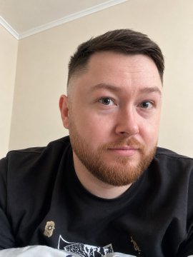A close-up selfie of a man with short brown hair and a reddish beard wearing a black graphic t-shirt, looking directly at the camera.