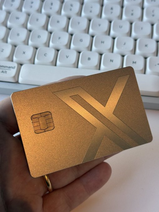 A hand holds a gold-colored metal card featuring a large "X" design and an EMV chip, displayed in front of a white keyboard. The card visually represents the "#Xmoney" hashtag from the post, showcasing what appears to be a premium branded payment or membership card for the X platform.