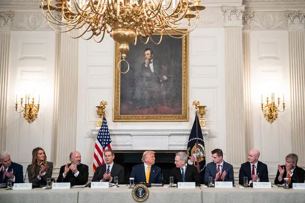 Officials including Donald Trump sit at a long table in a formal room with a chandelier and Lincoln portrait, clapping during a meeting.