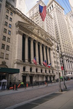 Street view of the New York Stock Exchange (NYSE) building with classical columns and American flags, located on a city street with surrounding tall buildings and a clear blue sky.