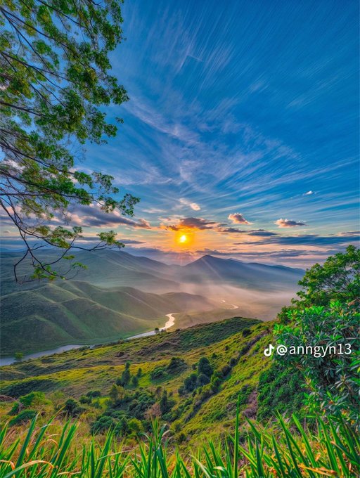 A scenic landscape photograph showing a sunset over a mountain range with a winding river, framed by tree branches, with a TikTok watermark '@anngyn13' in the corner.