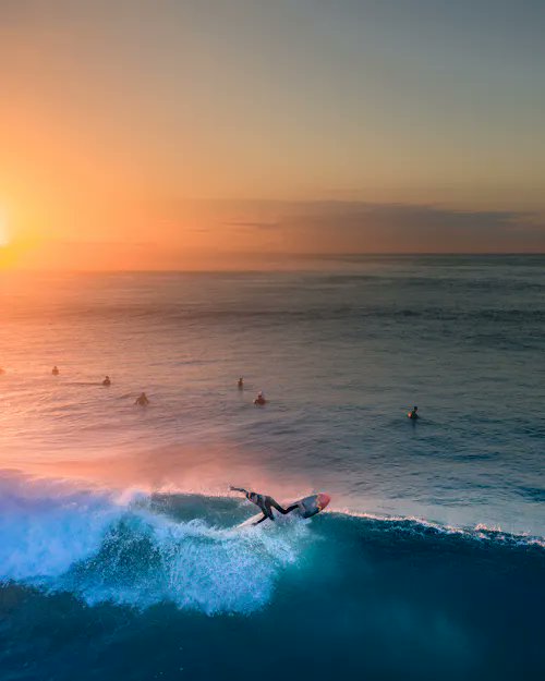A surfer carves through a glowing wave at sunset, with other surfers waiting in the distance as the sky blazes in warm orange hues. The image's radiant golden light perfectly matches the "Glow Show" theme of this NFT sale, which sold for 0.25 ETH ($616.18) as part of the $RARE collection.