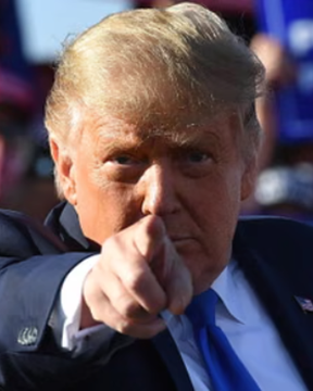 A close-up photograph of Donald Trump pointing his finger directly at the camera with a serious expression, wearing a dark suit and blue tie.
