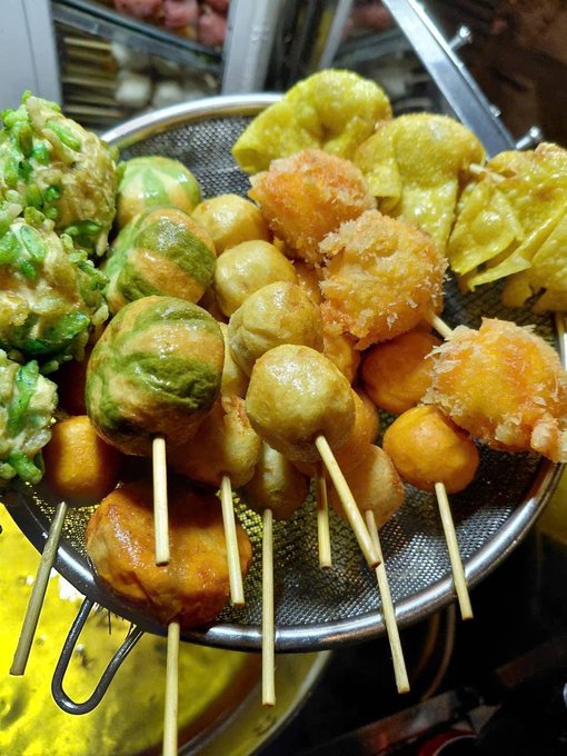 The image shows a silver wire mesh basket filled with various types of food, including fried balls, sticks, and other snacks. The basket appears to be placed on top of an oven or stovetop, suggesting that these items are being prepared for cooking or serving.