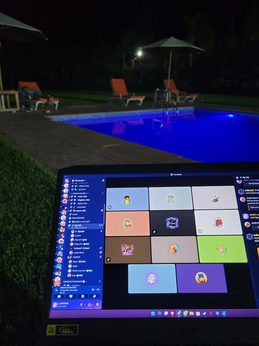 Laptop displaying a Discord call with multiple user avatars open on a desk by a swimming pool at night, with pool lounge chairs and umbrellas visible in the background.
