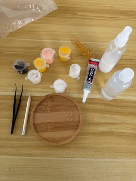 The image shows a wooden table with various items, including paint tubes, brushes, and other art supplies. The scene also includes a bottle of glue, which could be used for attaching different materials together or as an additional element in artwork. The presence of these items suggests that someone might have been working on a creative project involving painting and crafting.