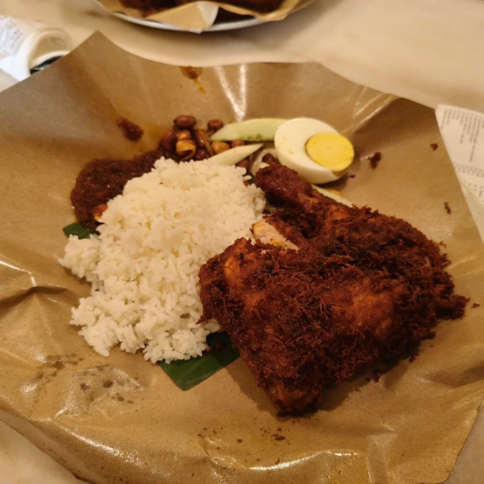 Nasi Lemak served on brown paper at Village Park in Malaysia, featuring coconut rice, a fried chicken leg with crispy crumbs, sambal, cucumber slices, and a half-boiled egg. The post highlights the dish as one of the best in Malaysia, noting the meal cost around 0.0014 ETH.