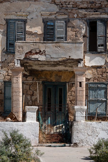 An old, weathered two-story building with peeling paint, broken windows, and blue shutters, featuring a gated entrance.