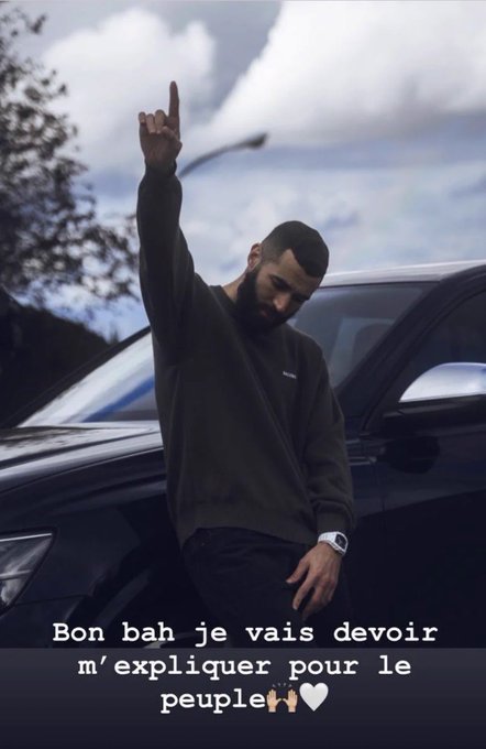 A man in a black sweatshirt leaning against a black car, raising his right index finger skyward, with French subtitles reading 'Bon bah je vais devoir m'expliquer pour le peuple'.
