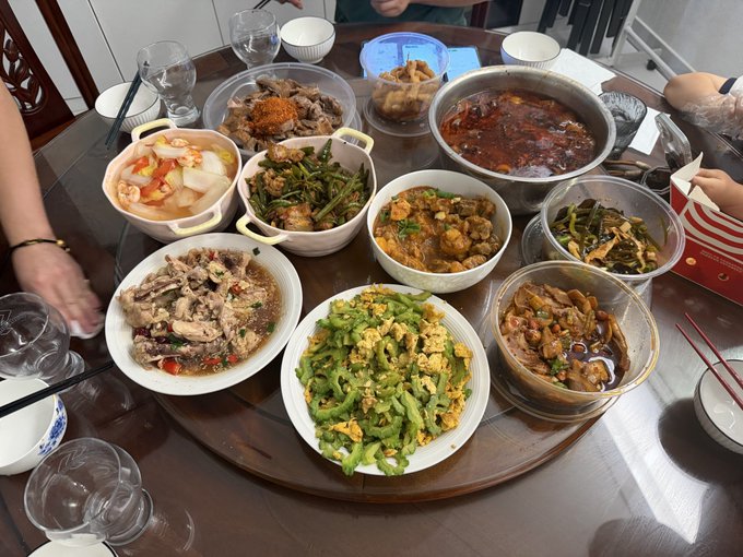 A photograph of a round dining table laden with a family-style Chinese meal featuring numerous dishes including bitter melon with egg, spicy stir-fried greens, meat dishes, and a hot pot, with people's hands and chopsticks visible around the table.
