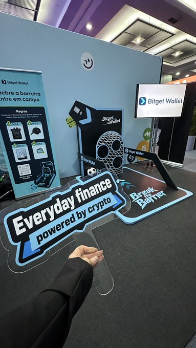 A hand holds a sign reading 'Everyday finance powered by crypto' in front of a Bitget Wallet promotional booth at an event.