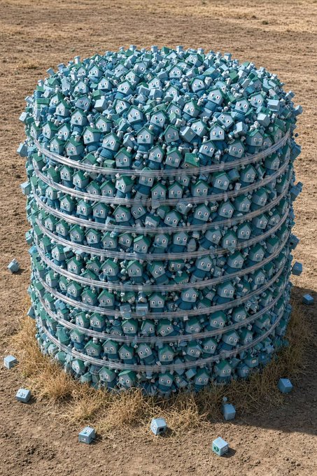 A surreal 3D render of a massive cylindrical pile of blue house-shaped objects stacked in layers on a dirt surface.