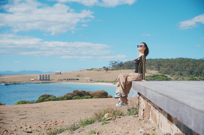 A woman sits on a stone ledge with her face tilted toward the sun, wearing sunglasses and a black-and-yellow striped top, overlooking a serene bay with rolling hills and scattered ruins in the distance. The image captures a peaceful, sun-drenched morning, matching the cheerful "GM～GMMMM～～～～" greeting.