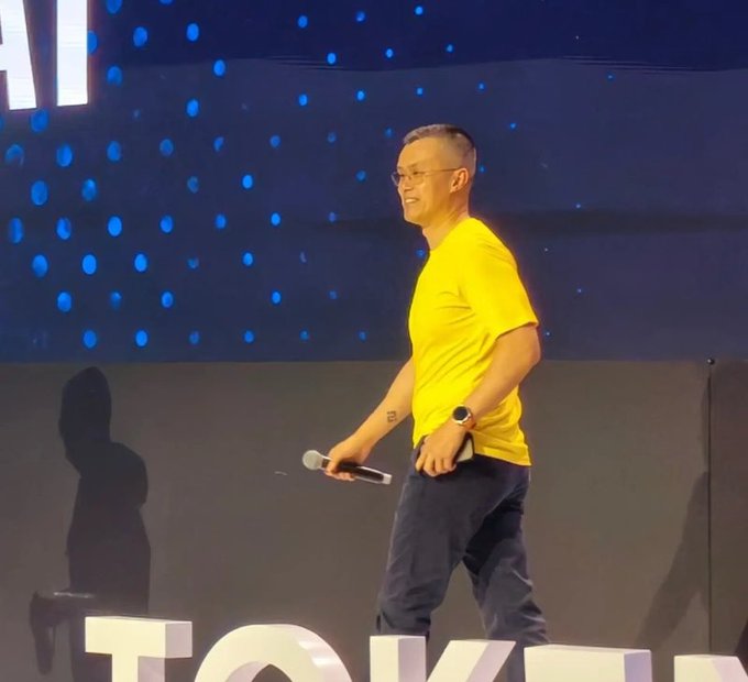 A man in a yellow t-shirt and dark pants holds a microphone while walking on a stage with blue dotted lighting and 'TOKEN' visible in the background.