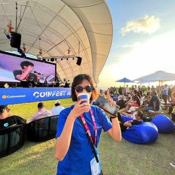 A man in a blue shirt and sunglasses speaks into a microphone while holding a smartphone at the CoinFest Asia outdoor event, with attendees relaxing on bean bags under a large white canopy.