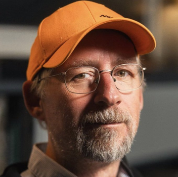 A close-up portrait of a bespectacled man with a gray beard wearing an orange baseball cap, looking to the side in a thoughtful expression — matching the caption's reference to the "cypherpunk legend" being featured. No visible text appears in the image itself.