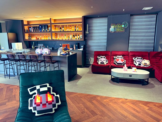 A modern lounge or hotel room featuring a bar area, plush red sofas, and distinctive black and white pixel-art cushions, with a neon 'To The Moon' sign glowing on the wall.