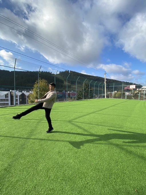 A man in a beige jacket and black pants performs a high kick on a bright green artificial turf soccer field, with hills and a fence visible in the background.