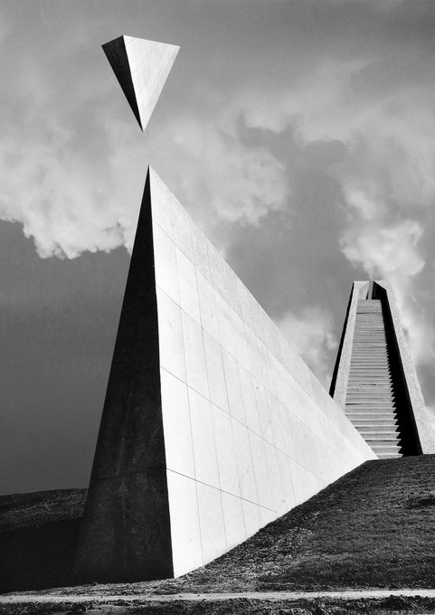 A black and white photograph of a modern, angular architectural structure with a triangular wedge seemingly floating in the sky above a pyramidal tower with a staircase, set against a cloudy backdrop.