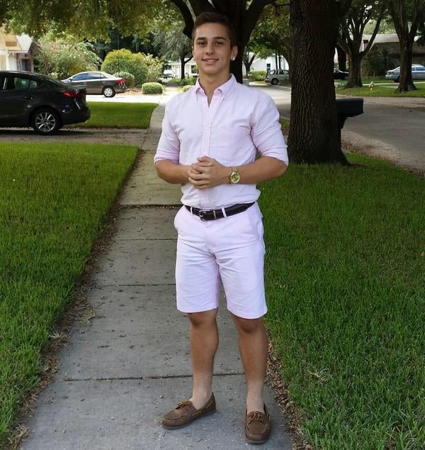 A photograph of a young man wearing a light pink button-up shirt, pink shorts, a black belt, and brown loafers, standing on a sidewalk in a suburban neighborhood.