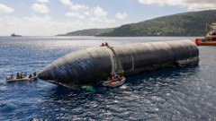 An aerial view of a large black inflatable object being towed or moved by small boats in the water near a coastline.
