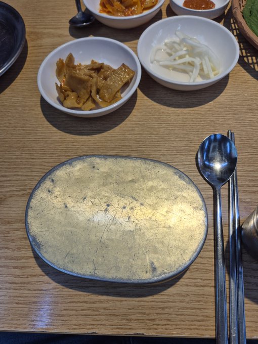 A Korean restaurant table setting featuring an empty gold-patterned plate, a spoon and chopsticks, and side dishes including pickled radish and seasoned vegetables.