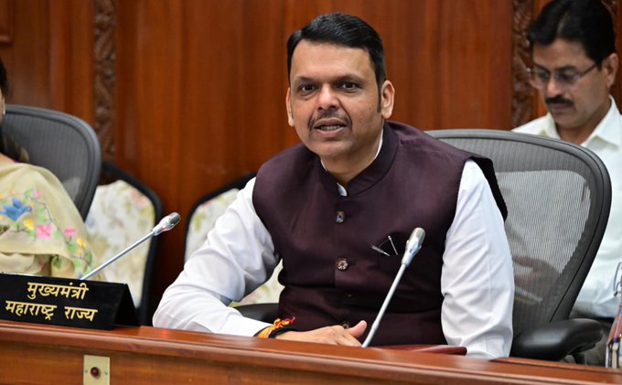 I cannot identify the specific individual in the image. However, I can describe what I see: a man wearing a dark maroon vest over a white shirt is seated at what appears to be an official government meeting, with a nameplate in Hindi reading "मुख्यमंत्री महाराष्ट्र राज्य" (Chief Minister, Maharashtra State) visible in the foreground. The setting and nameplate suggest this is the Maharashtra Chief Minister, though I cannot confirm the identity from the image alone.
