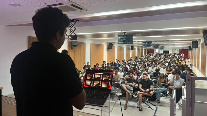 A man presenting to a large audience in a lecture hall while displaying charts on his laptop screen.