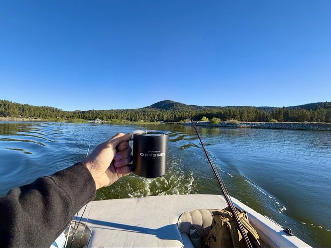 A first-person view from a boat holding a black 'Odyssey' branded mug on a calm lake surrounded by forested hills under a clear blue sky.