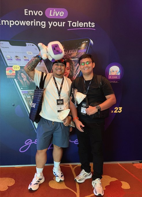 Two men pose in front of an "Envo Live - Empowering your Talents" banner at what appears to be a convention, one holding a promotional plush toy.
