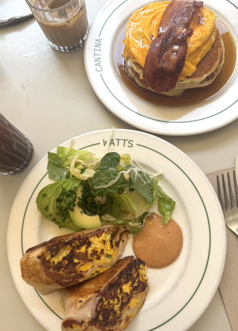 Two plates from Cantina Watts display a mouthwatering breakfast: pancakes topped with a cheesy omelet and crispy bacon drizzled in syrup, alongside a side salad, avocado, and cheesy hash browns, paired with a latte—worth every bit of the 27 $KAITO paid to celebrate a boyfriend's birthday.