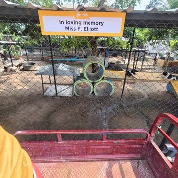 A photograph taken from the back of a red truck showing a memorial site enclosed by a chain-link fence, featuring a yellow sign reading 'In loving memory Miss F. Elliott' above stacked concrete rings used for an enclosure.