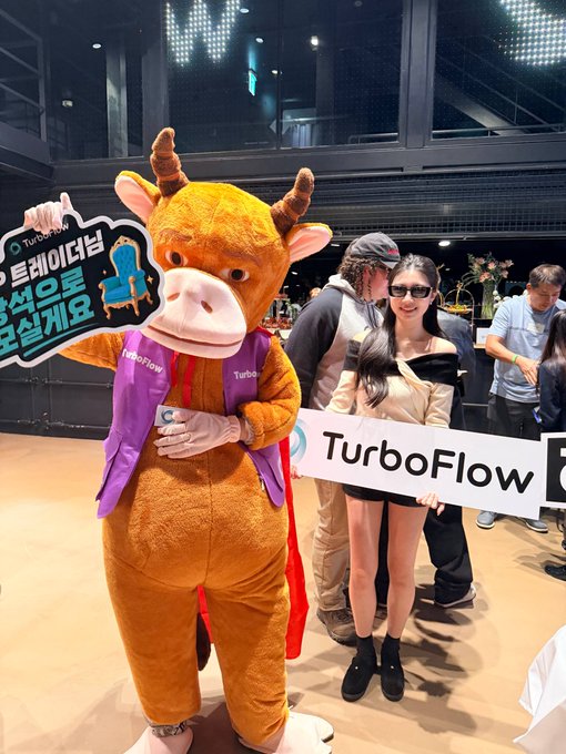 A person in a cow mascot costume promoting TurboFlow at an event, holding a sign in Korean asking visitors to sit on the chair, while a woman holds a 'TurboFlow' banner.