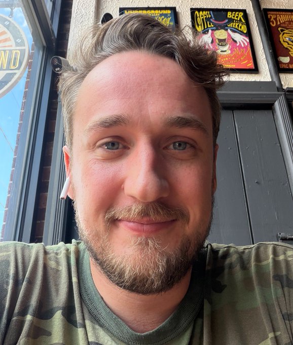 A selfie of a smiling man with short brown hair and a beard, wearing AirPods and a camouflage shirt, taken outside a building with beer-related signs visible in the background.