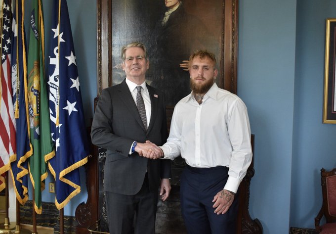 Two men shaking hands in a formal government room with American and other flags and a portrait in the background; one wears a dark suit and the other is Jake Paul in a white shirt.