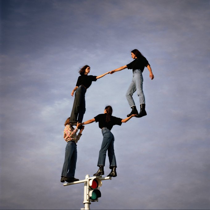 A surreal photograph of three women in black tops and jeans balancing on a traffic light, holding hands in a human pyramid formation against a cloudy sky.