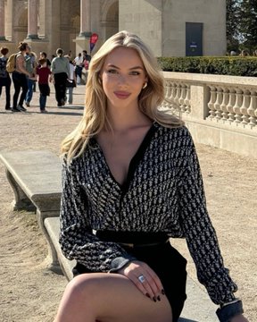 A photograph of a blonde woman sitting on a stone bench outdoors, wearing a black and white patterned blouse with a black skirt.