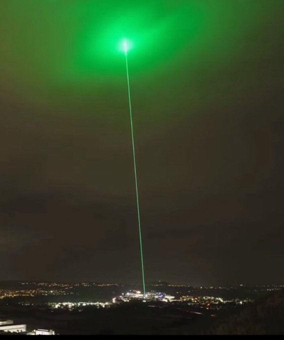 A nighttime scene featuring a powerful green laser beam shooting straight up into the sky from a city below, creating a bright glowing halo effect at its peak.