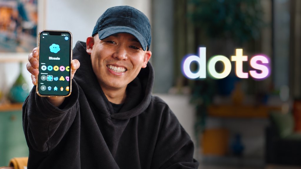 A smiling man wearing a cap and dark hoodie holds up a smartphone displaying an app called 'Blosso' with various colorful icons, while a glowing 'dots' logo is visible in the background.