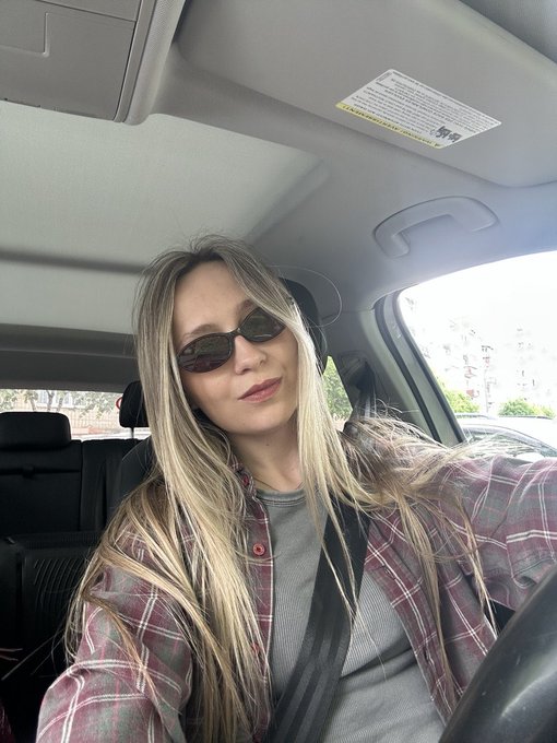 A blonde woman wearing sunglasses, a gray top, and a plaid shirt takes a selfie from the driver's seat of a car.