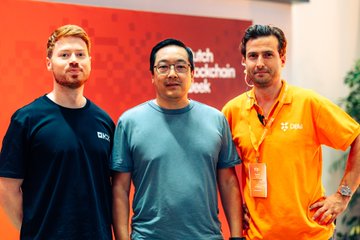Three men posing together in front of an orange "Dutch Blockchain Week" backdrop, wearing a dark SQX shirt, blue tee, and orange polo respectively.