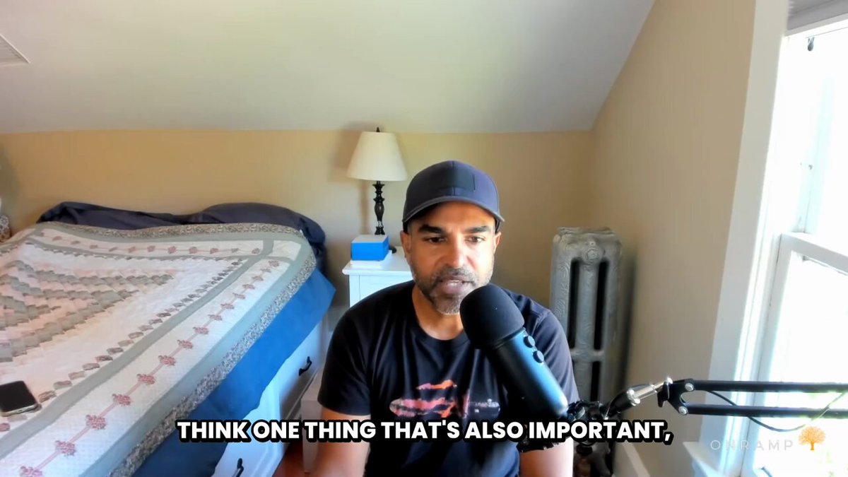 The image shows a man sitting at a desk with a microphone, wearing a black shirt and hat. The background includes a bed, lamp, and window. There are also two texts displayed in the image that read "Think one thing that's actually important" and "$MSTR | $STRC | $SATA".