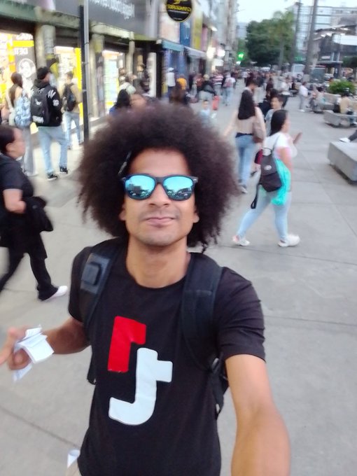 A selfie of a smiling young man with an afro hairstyle wearing blue reflective sunglasses and a black t-shirt with red and white graphics, standing on a busy pedestrian street.