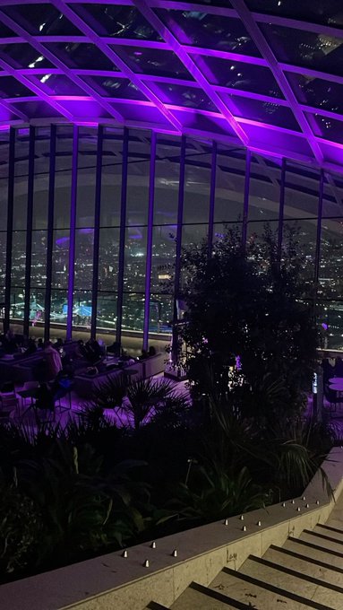 An indoor lounge area features floor-to-ceiling windows overlooking a city skyline at night, illuminated by purple ceiling lights.