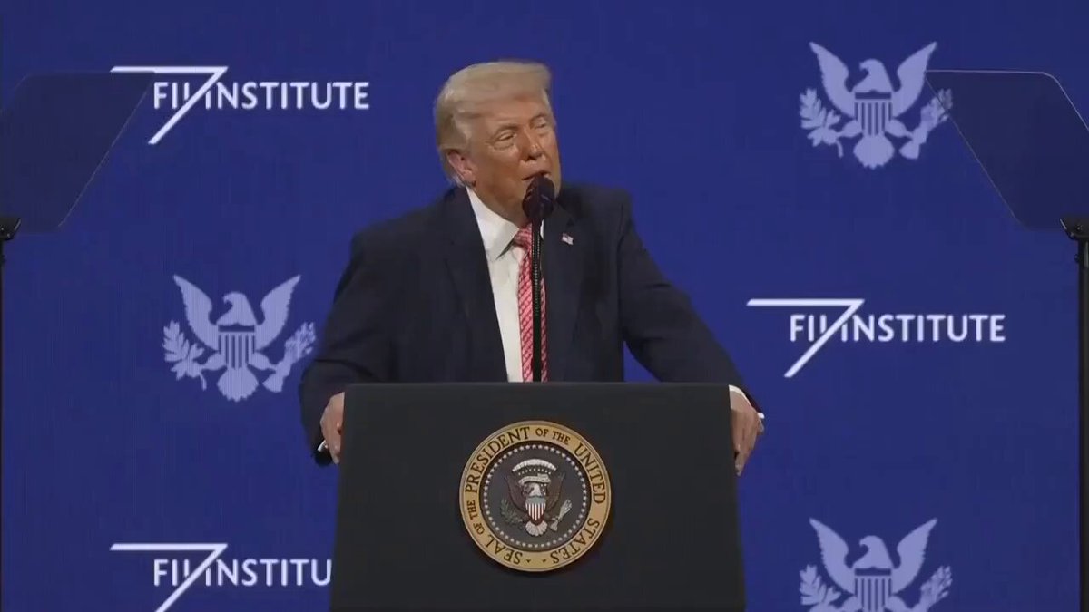 The image features a man standing at a podium, giving a speech or addressing an audience. The man appears to be wearing a suit and tie, suggesting that he might be a political figure such as President Trump. He is speaking into a microphone, which can be seen in front of him on the stage. The backdrop of the scene includes a blue wall with various logos, including one for "Founded Institute" and another for "The Adoption Wave". This suggests that the event could be related to cryptocurrency or digital assets, as Trump has been advocating for their adoption by world leaders.