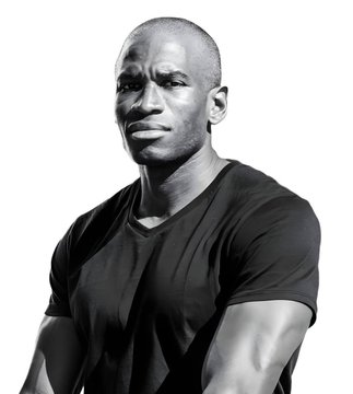 A black and white portrait of a muscular, bald man wearing a black V-neck t-shirt, posing with his arms crossed.