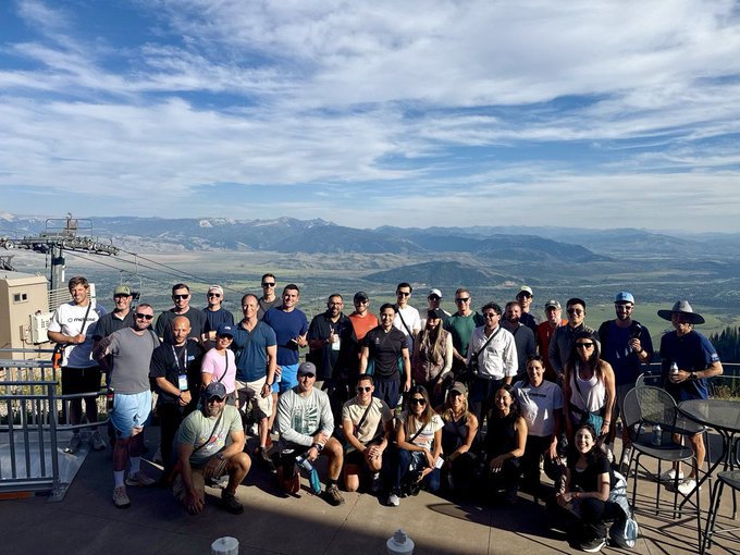 A large group photo of approximately 30 people posing together on a mountain observation deck, with a stunning view of green valleys and mountain ranges in the background under a partly cloudy sky.