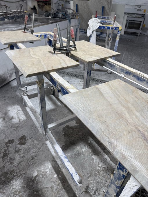 A photograph of stone or marble slabs resting on metal saw stands in a workshop, with wet floors and industrial equipment visible in the background.