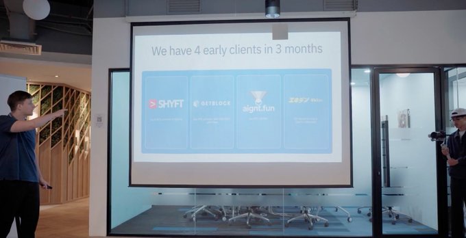 The image shows two men standing next to a large screen displaying information about a company called "rapid8n". One man is pointing towards the screen, while another man is holding a camera, possibly capturing the moment or taking notes on the presentation. The screen displays four different logos of companies, including "rapid8n", "solana", and "snoop".