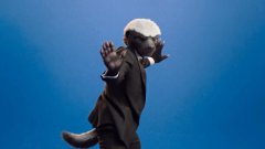 A 3D-rendered anthropomorphic rat or mouse character wearing a brown robe, gesturing with its hands against a clear blue sky.